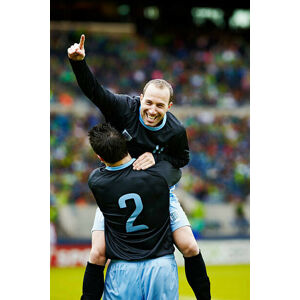 Umělecká fotografie Soccer player jumping into the arms of teammate, Thomas Barwick, (26.7 x 40 cm)